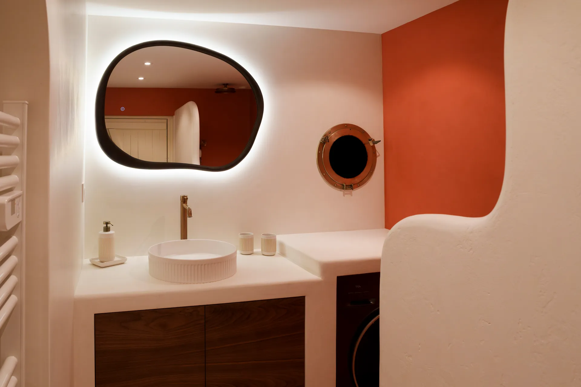Salle de bain béton ciré — vasque, miroir organique, hublot et mur terracotta, vue d'ensemble